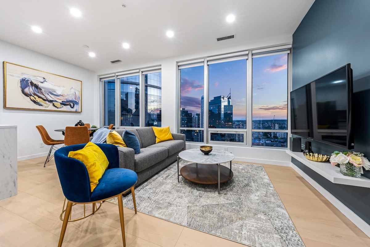 A modern living area features a spacious sectional sofa with decorative yellow pillows. A contemporary coffee table sits on a soft area rug, while large windows showcase a vibrant city skyline and sunset views. A wall-mounted television is positioned across from the seating area.