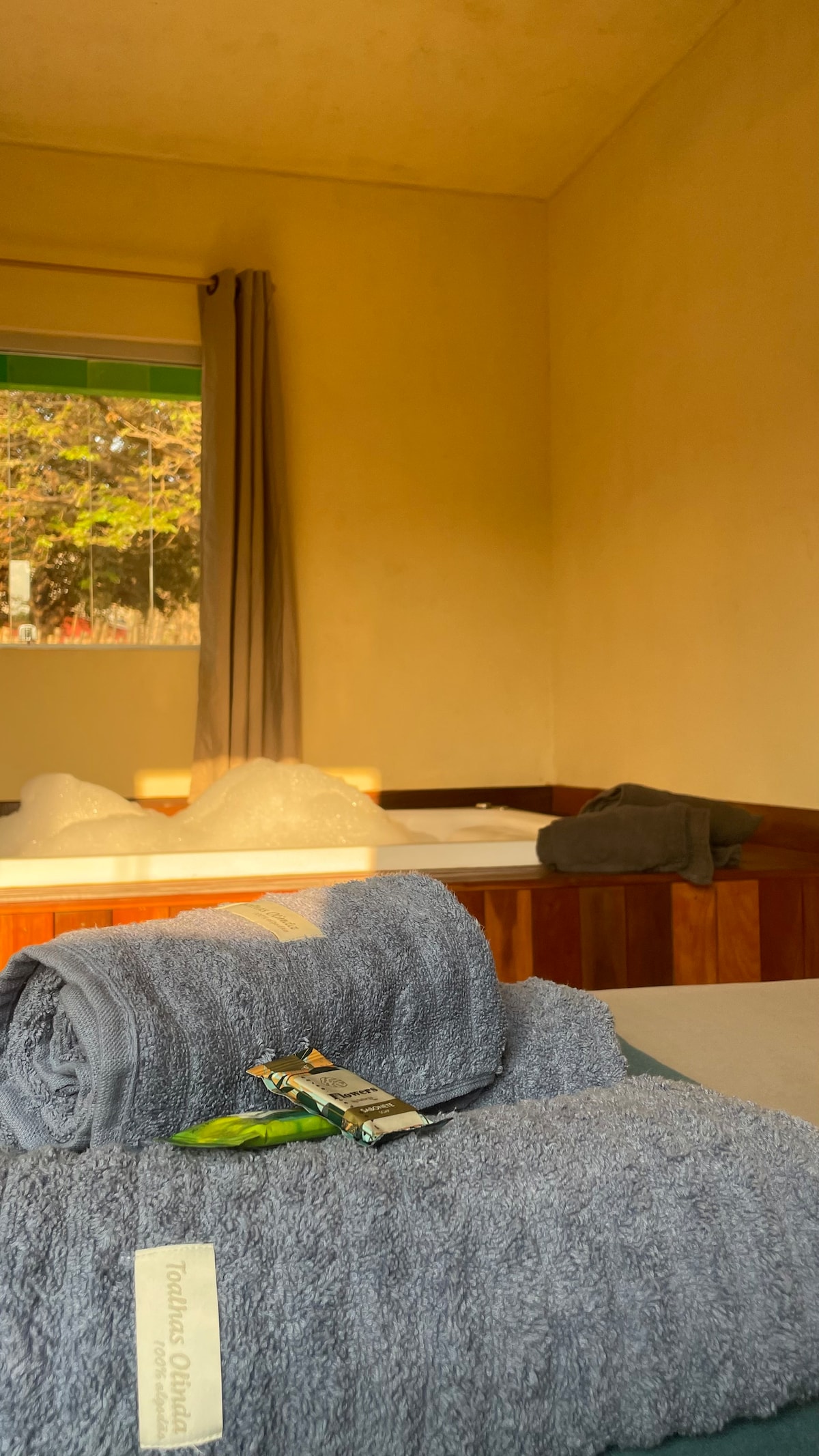 A well-lit bedroom features soft towels neatly placed on the bed, accompanied by small toiletries. Light filters through a window adorned with light curtains, illuminating the warm-toned walls and wooden accents throughout the space.