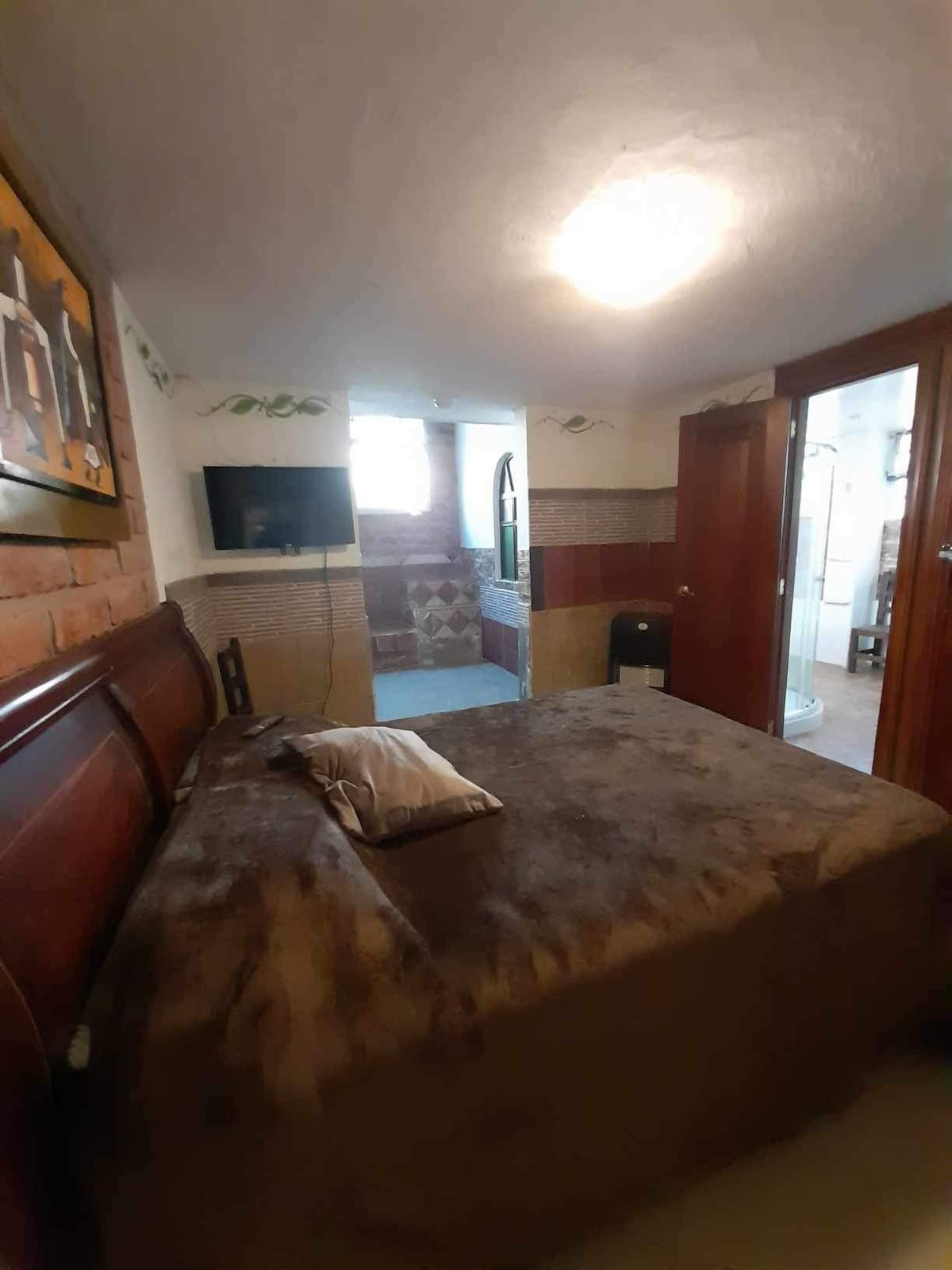 A cozy bedroom features a spacious bed covered with a soft brown blanket and a decorative pillow. The room includes a mounted television and is well-lit by a nearby window. Access to a bathroom is visible through a wooden door.