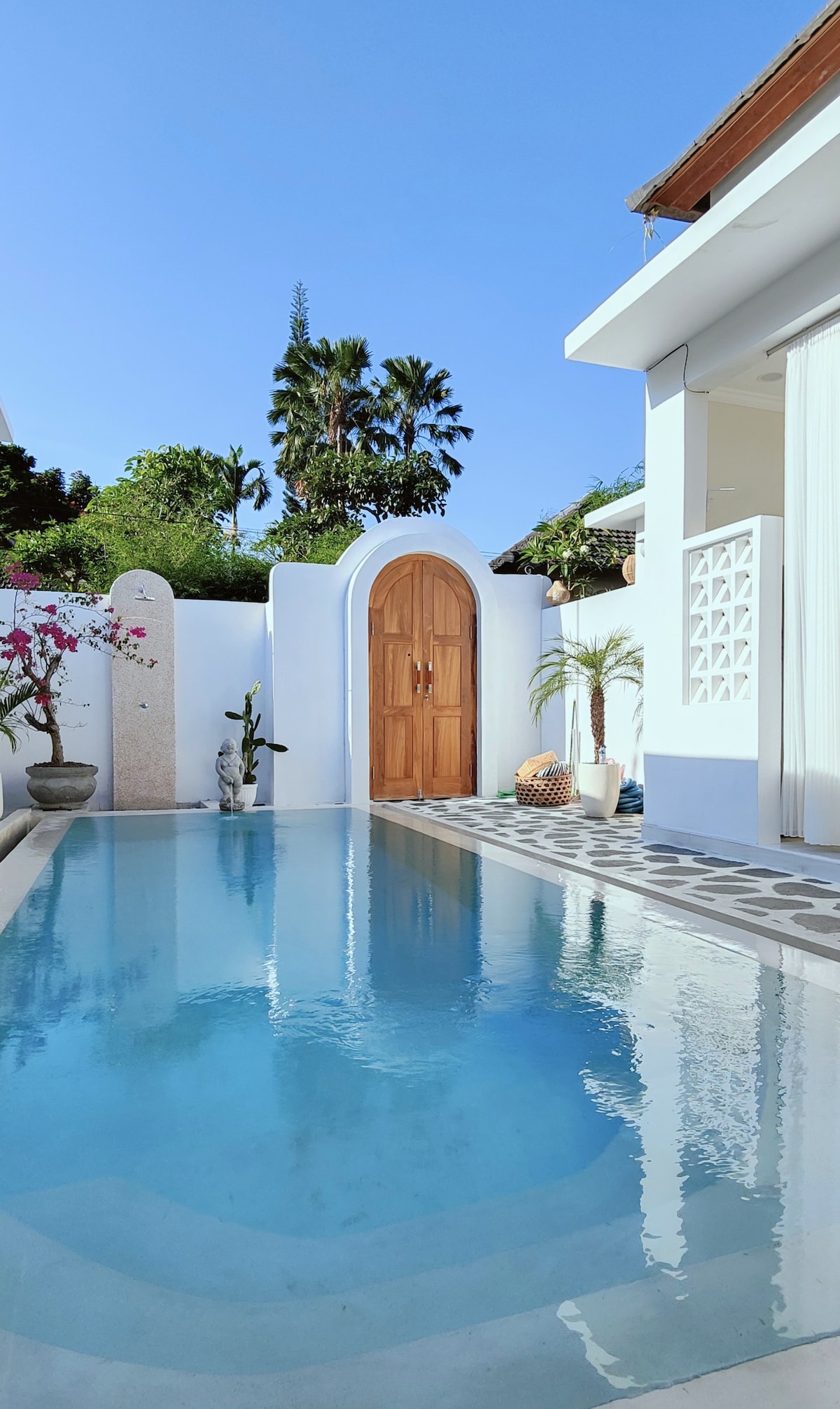 A private swimming pool reflects the blue sky, framed by tropical greenery and a wooden door leading to the villa. The surrounding area features patterned tiles and potted plants, creating a serene outdoor space for relaxation.