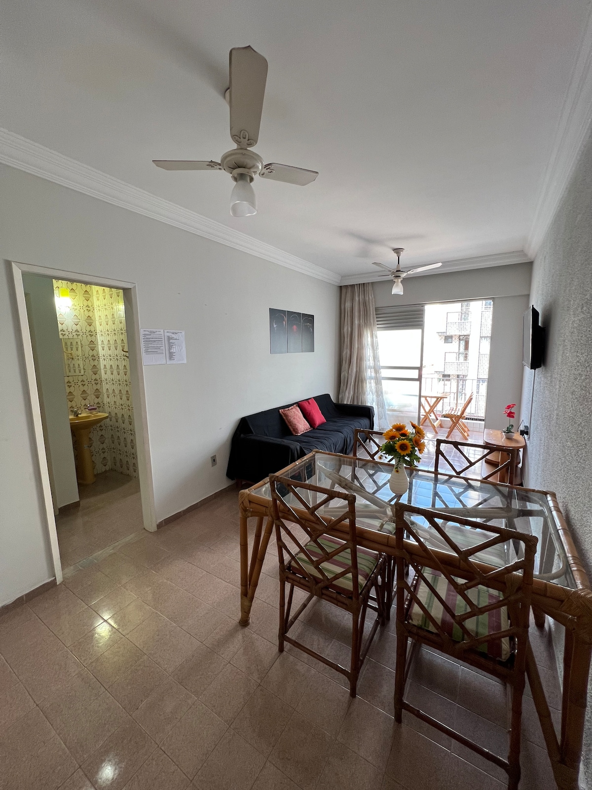 A spacious living area is shown, featuring a light-colored wall and a comfortable black sofa. A glass dining table with four bamboo chairs is visible, complemented by a vase of flowers. A doorway in the background leads to a bathroom with tiled walls.