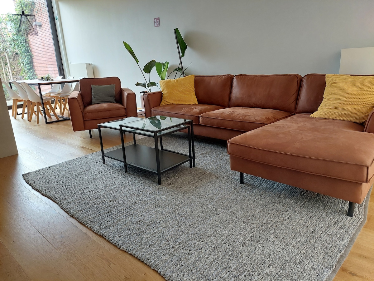 A spacious living area features a brown leather sectional sofa complemented by two yellow cushions. A glass coffee table rests on a textured gray rug. In the background, a dining table with light-colored chairs is visible, alongside large windows providing ample natural light.