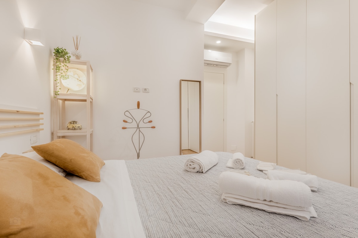 A spacious bedroom features a large bed dressed in neutral bedding, complemented by two decorative pillows. Soft towels are neatly arranged at the foot of the bed. A modern wardrobe and a small shelf with decorative items are visible, enhancing the overall light and airy ambiance.