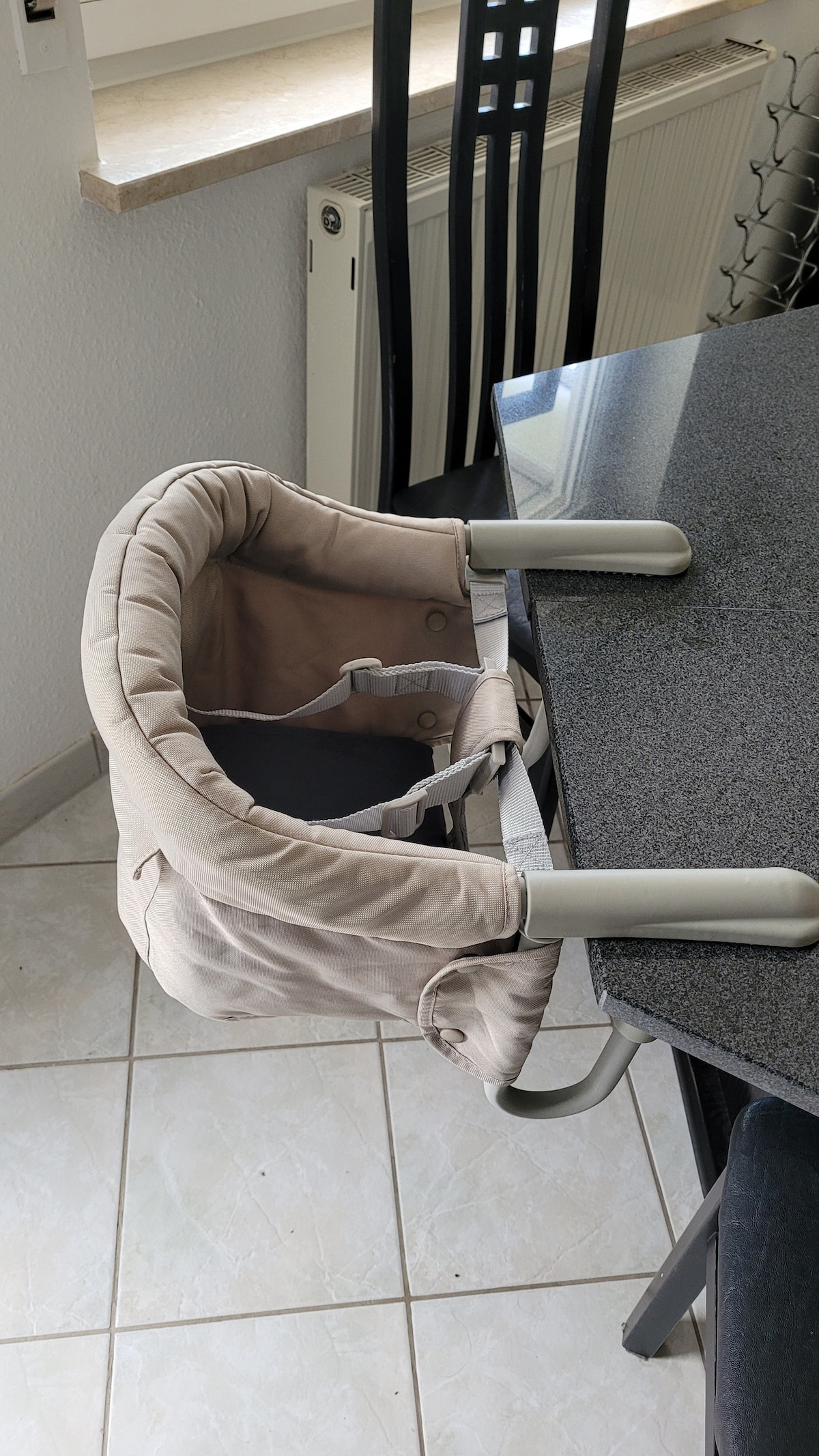 A high chair is shown attached to a sleek, dark granite kitchen table. The chair features a cushioned seat and safety straps, ensuring comfort and security for young children. Light reflects softly on the surrounding tile flooring and table surface.