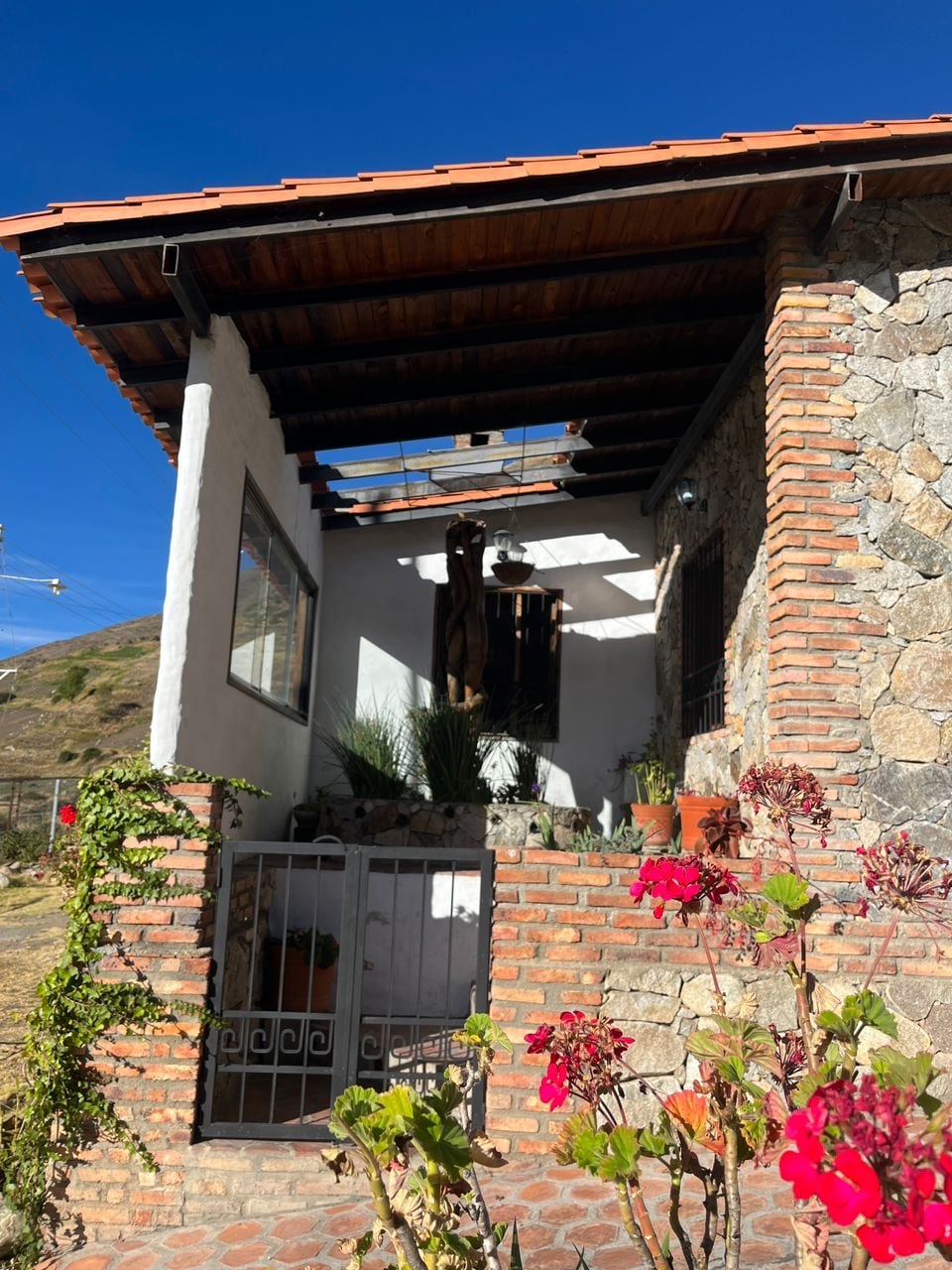 The entrance showcases a charming mix of stone and brick, framed by vibrant flowers and greenery. A wooden overhang offers shade, while large windows allow for natural light. Decorative elements such as potted plants enhance the inviting atmosphere of this mountain getaway.