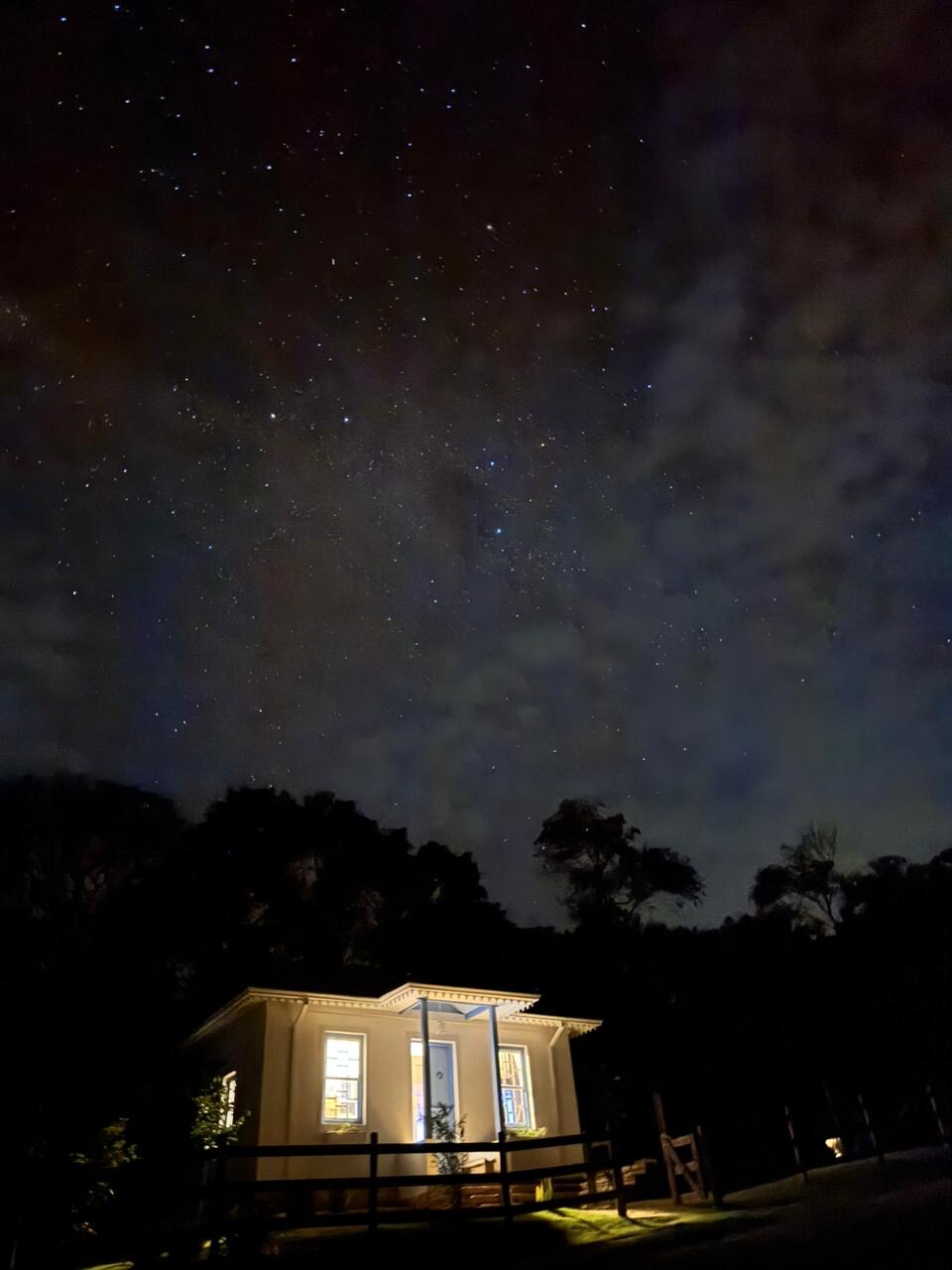 The image captures a charming colonial-style house illuminated against a night sky filled with stars. The gentle glow from the windows hints at a welcoming interior, while dark silhouettes of surrounding trees frame the scene, emphasizing the tranquil, rural atmosphere.