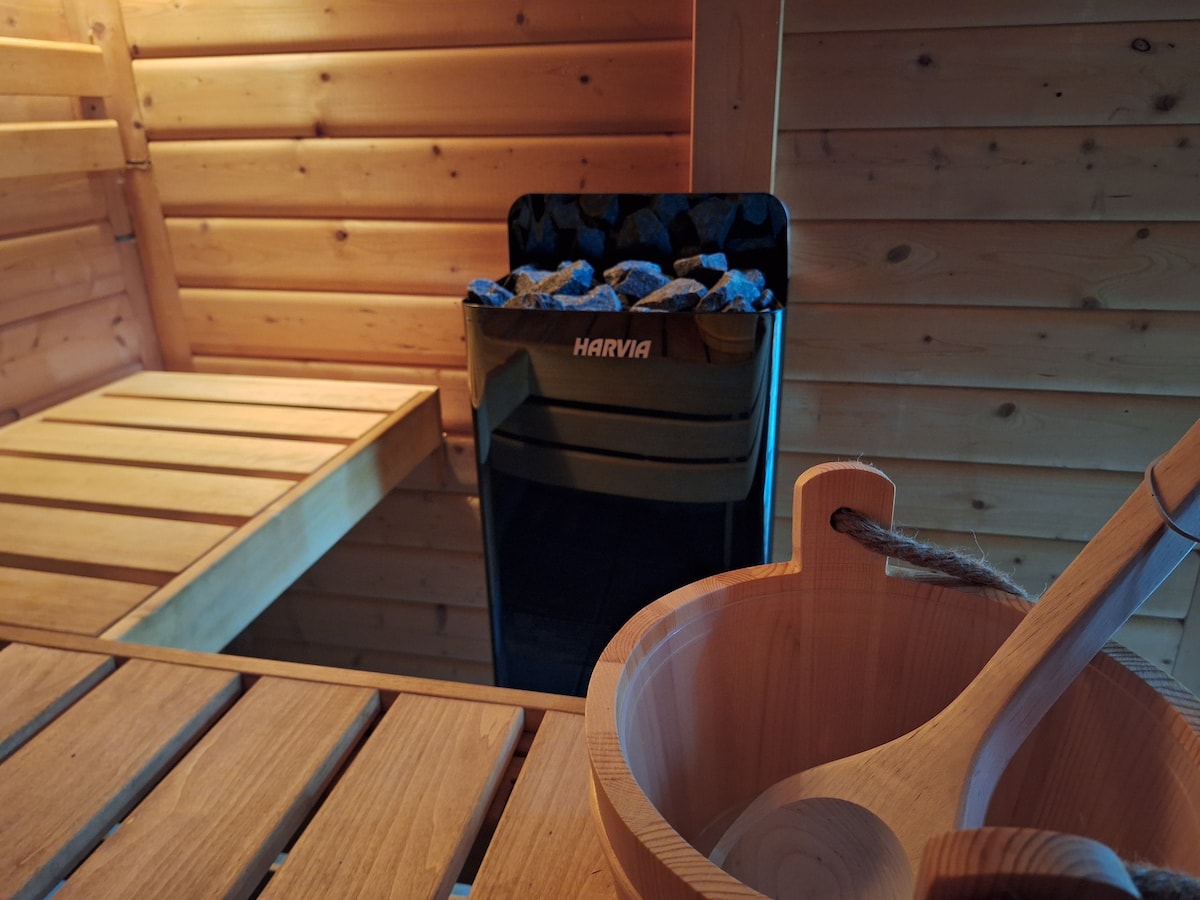 A private sauna is captured, featuring warm wooden walls and benches. A heater stands prominently with stones visible, while a wooden bowl and ladle rest beside a smooth wooden surface. Soft lighting creates a serene atmosphere, inviting relaxation.