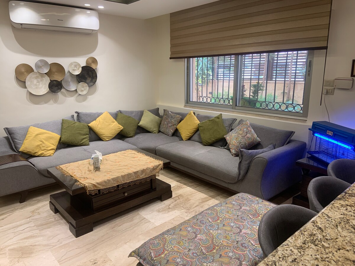 A spacious living area features a large, U-shaped gray sofa adorned with green and yellow cushions. A wooden coffee table sits at the center, complemented by a decorative textile. Natural light streams through large windows covered with blinds, creating a welcoming ambiance.