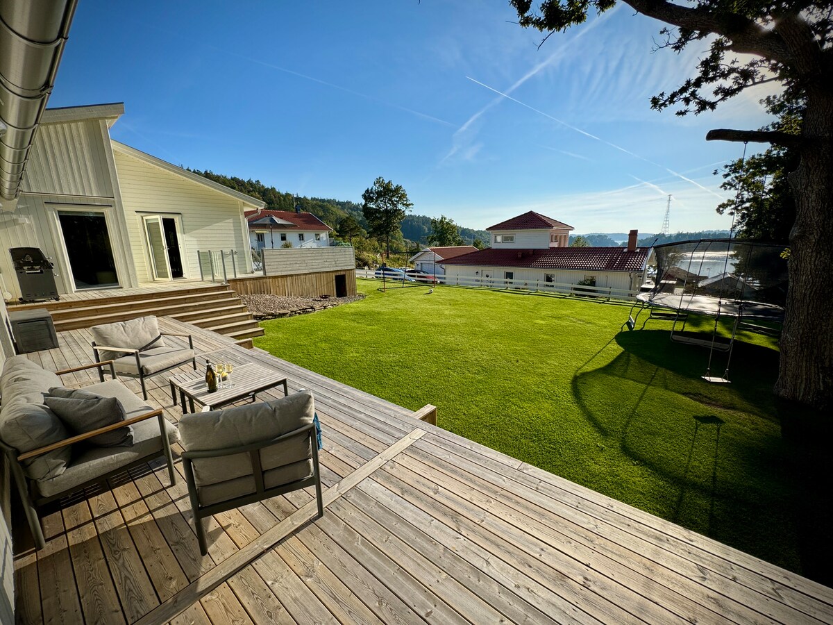 An outdoor seating area is presented on a wooden deck, featuring two chairs and a small table. The lawn is expansive and neatly maintained, complemented by a trampoline in the background. The surrounding area includes residential buildings and a clear blue sky overhead.