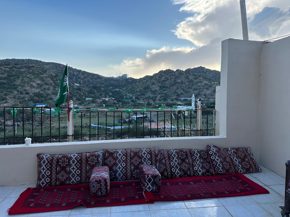 An outdoor seating area is highlighted, featuring low cushions arranged along a wall on a tiled floor. A traditional patterned rug adds a touch of warmth, while the view of rolling hills and valleys is visible in the background under a cloudy sky.