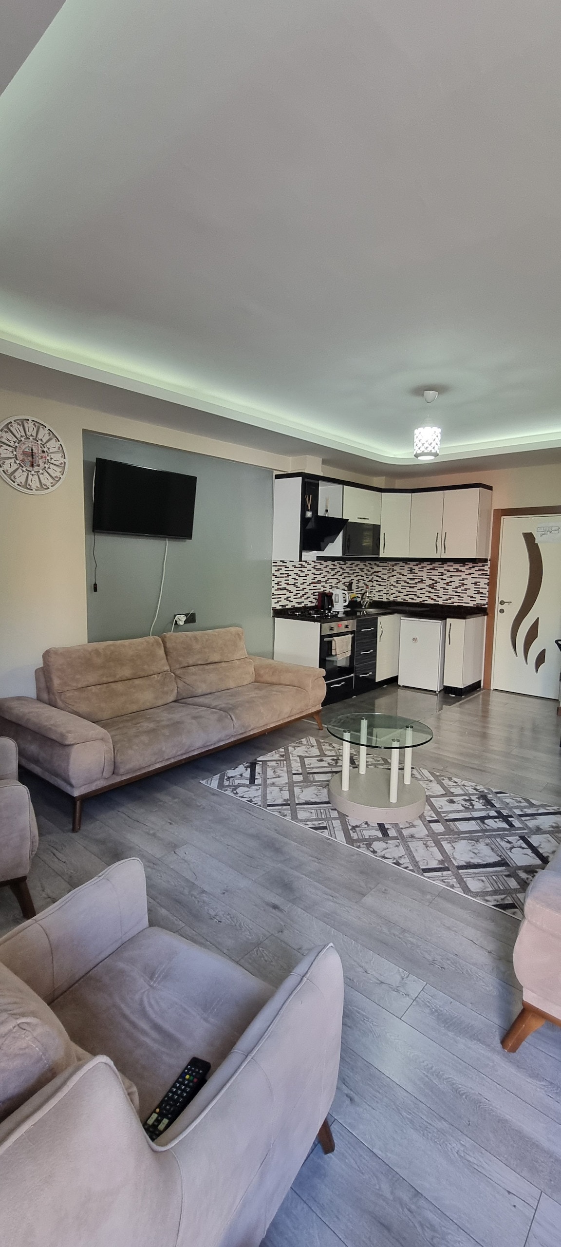 A modern living area is presented with a light-colored sofa set and a glass coffee table. The kitchen features contemporary cabinetry and appliances, while a television is mounted on the wall. Soft lighting is provided by a ceiling fixture, creating a welcoming ambiance.