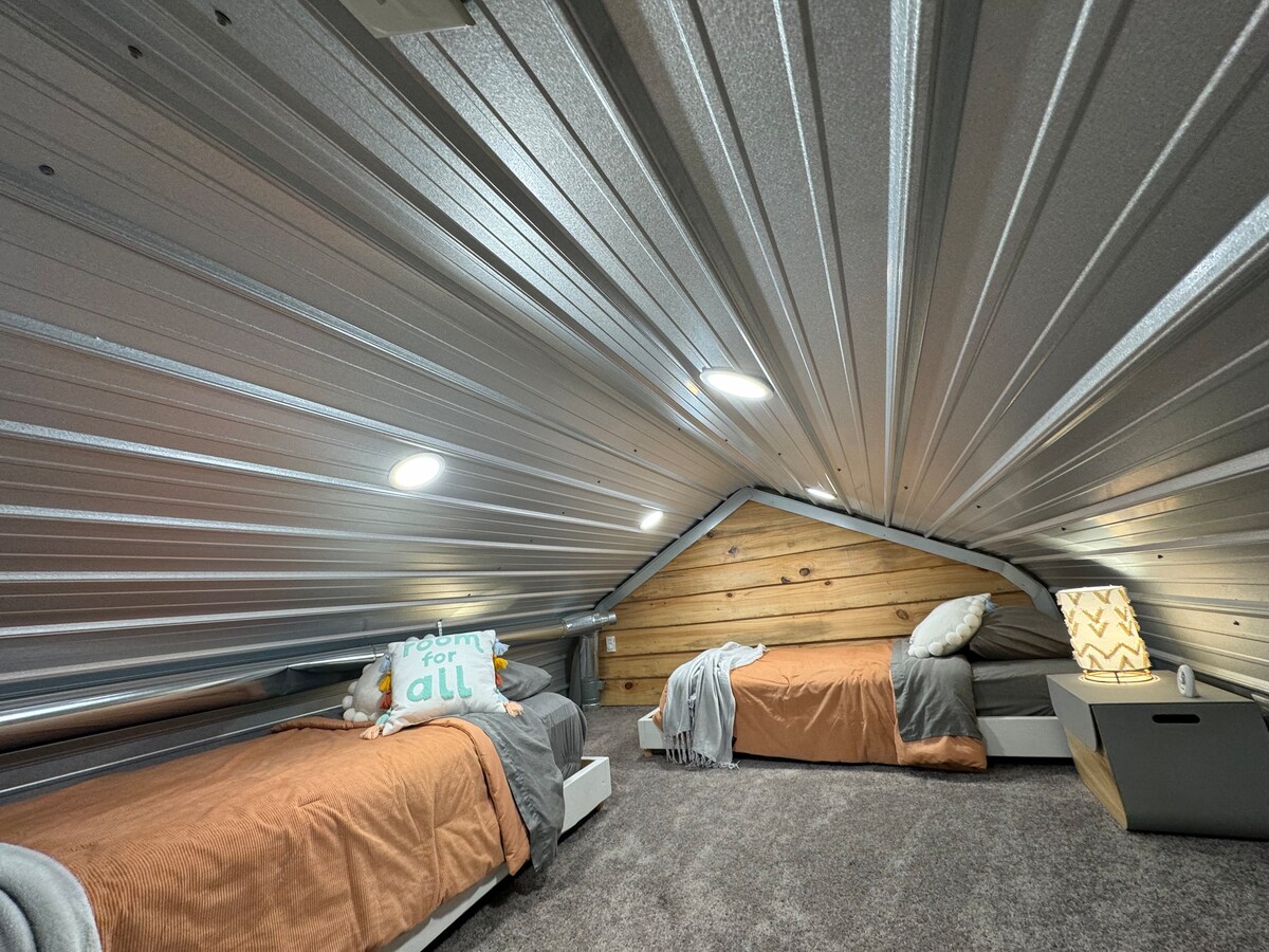 An attic space features two twin beds covered with neutral-toned blankets. Cozy pillows, including one with an inclusive message, are placed on the beds. The room is illuminated by recessed lighting in the sloped metal ceiling, and wooden paneling adds warmth to the decor.