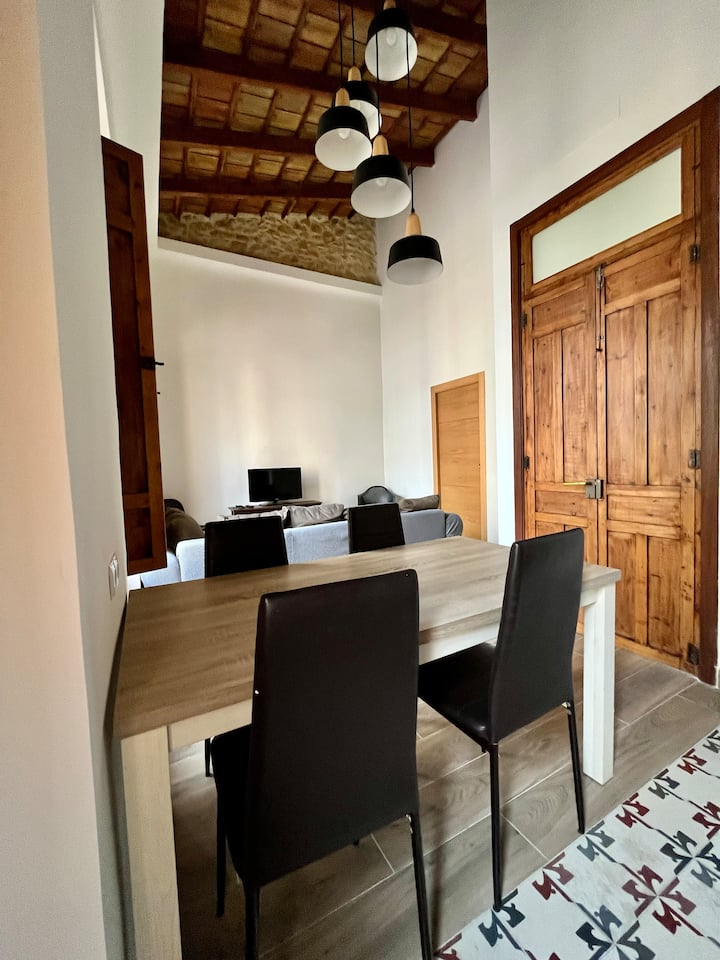 Espacioso salón comedor que destaca por sus techos altos con vigas de madera y su imponente puerta de entrada de madera noble. Un ambiente acogedor iluminado por lámparas de diseño moderno que combina historia y confort.