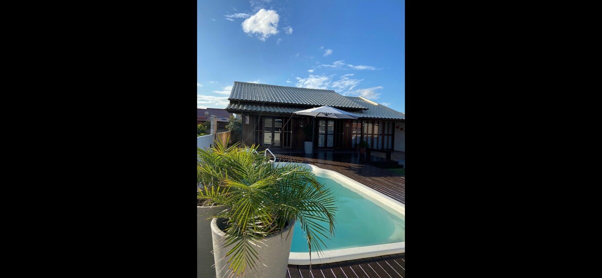 The exterior of the house is showcased, featuring a clear blue sky and a lush green palm in the foreground. A private pool is visible, with a shaded area provided by an umbrella. The house has a relaxed architectural style with large windows and a welcoming entrance.