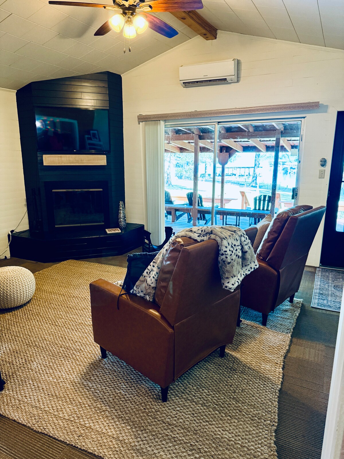 A cozy living area features two plush, brown armchairs positioned around a neutral area rug. A large window allows natural light to fill the space, showcasing views of the outdoor patio. A subtle fireplace is located on the opposite wall, contributing to a welcoming environment.