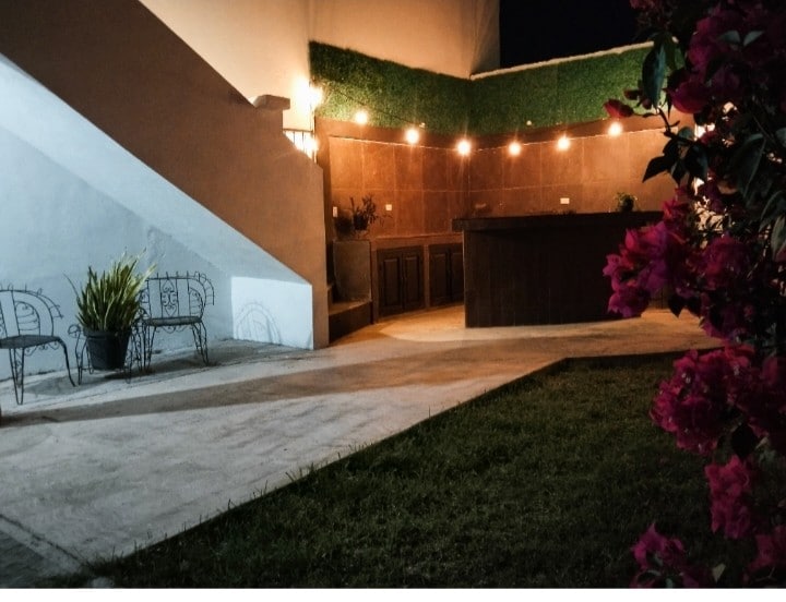 An outdoor space is showcased, featuring a pathway leading to a wooden bar area illuminated by subtle lighting. Two metal chairs are positioned beside a patch of grass, with lush plants bordering the area, creating a relaxed environment suitable for gatherings.