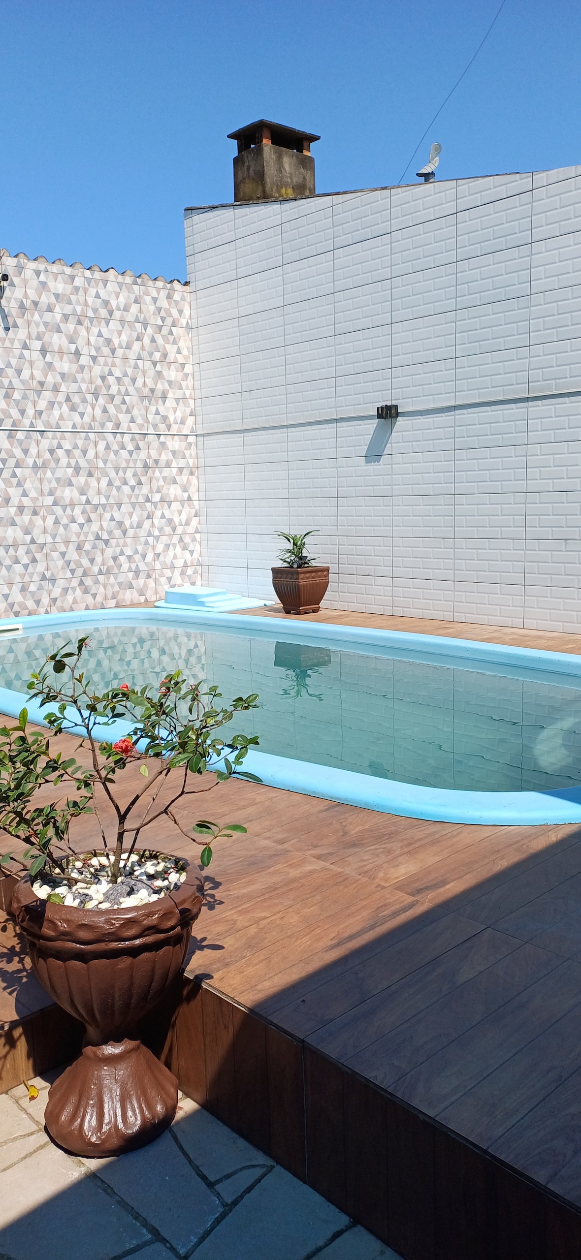 A private swimming pool is surrounded by a wooden deck, featuring two ornamental pots with greenery. The pool area is well-lit, showcasing clear water under a blue sky. The walls are accented with decorative tiles, enhancing the outdoor space.