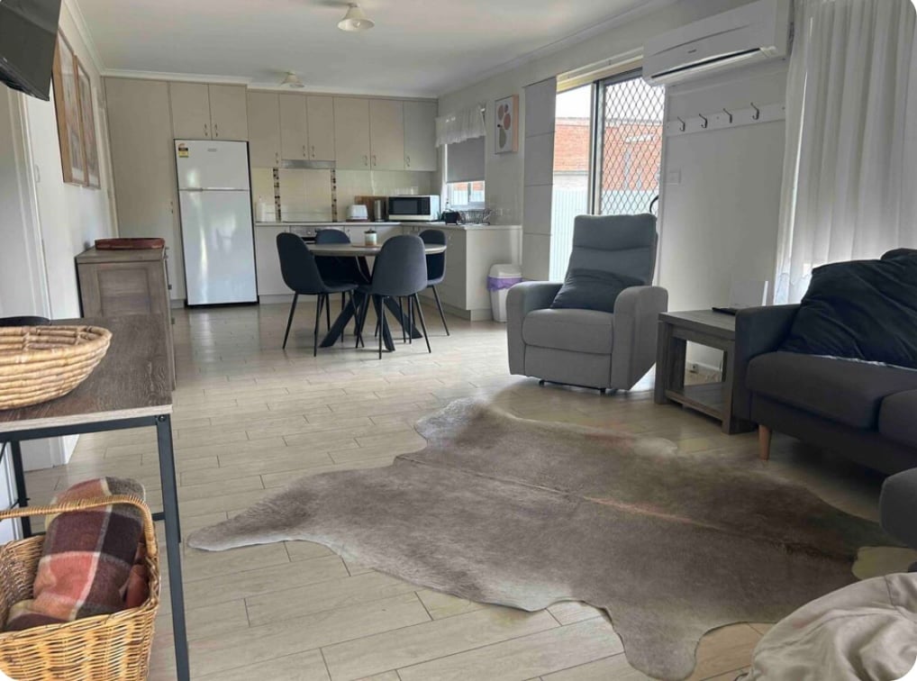 A spacious living area features a comfortable seating arrangement, including two gray armchairs and a sofa. A dining table with four chairs is visible, alongside a fully equipped kitchen with white cabinets. Natural light enters through the window, illuminating the room's neutral tones.