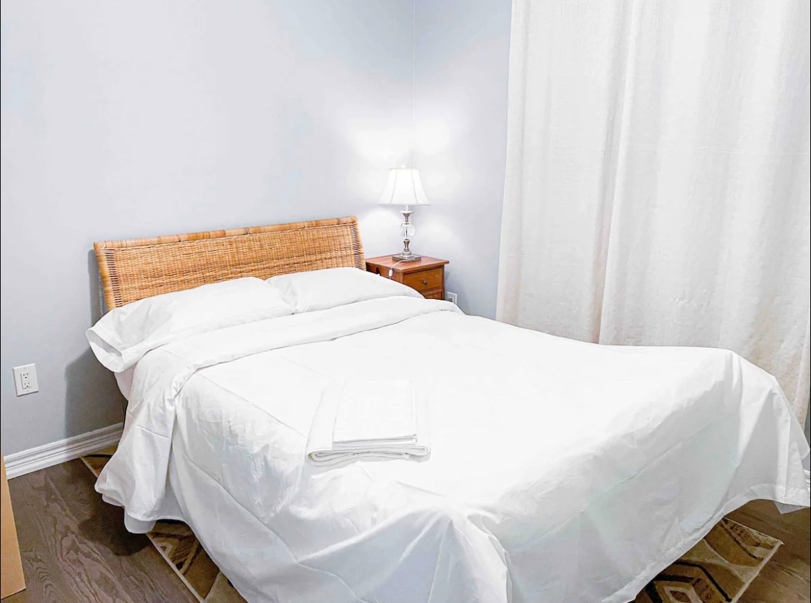 A well-lit bedroom features a spacious bed with crisp white linens, complemented by a woven headboard. A small side table with a lamp is positioned beside the bed. Light-colored curtains frame the window, enhancing the bright and airy atmosphere of the room.