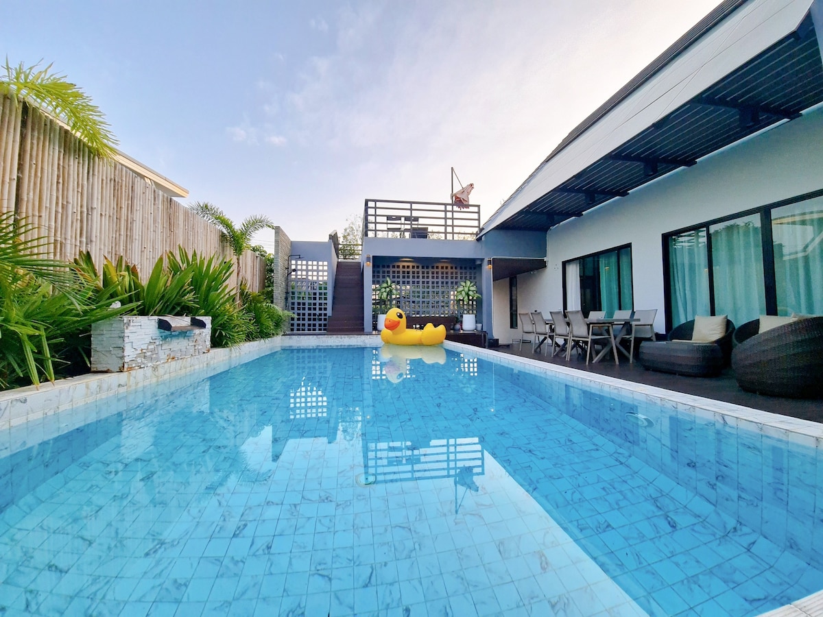 A private swimming pool is framed by lush greenery and a wooden fence. Sunlight reflects off the clear water, revealing a floating rubber duck. Patio furniture and a staircase are visible along the poolside, enhancing the outdoor relaxation space.
