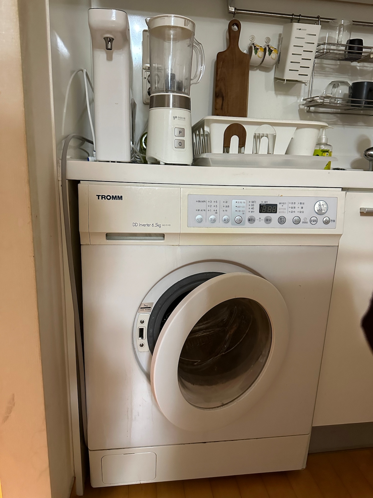 A white washing machine is positioned beside a blender on a kitchen counter. Above, various utensils and a wooden board are organized, while additional kitchen items are neatly arranged on shelves. The overall setup appears functional and well-maintained.