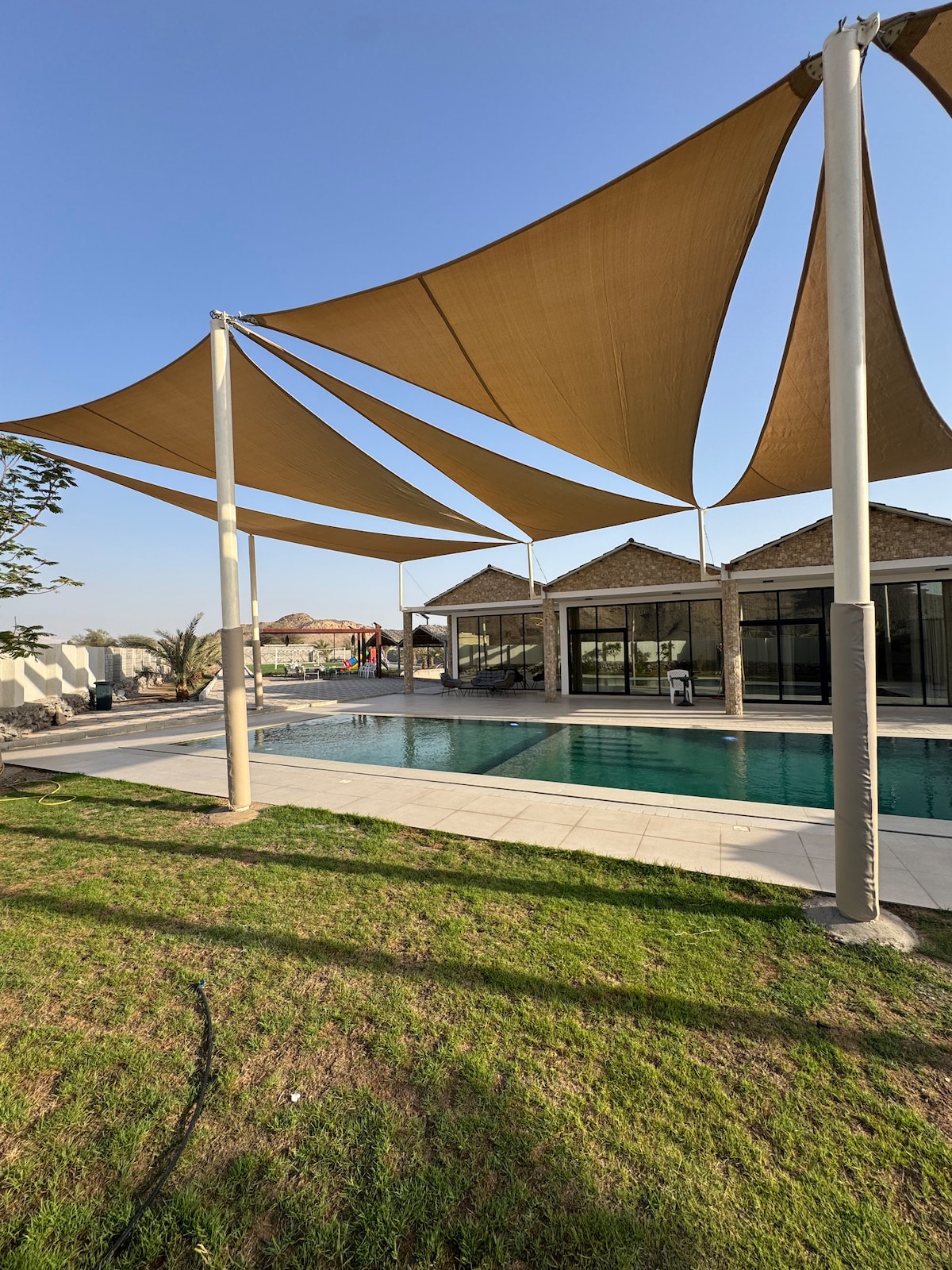 A shaded area is provided by large fabric canopies above the poolside, offering relief from the sun. The tranquil pool, surrounded by a tiled deck, reflects the blue sky and is complemented by well-maintained grass in the foreground.