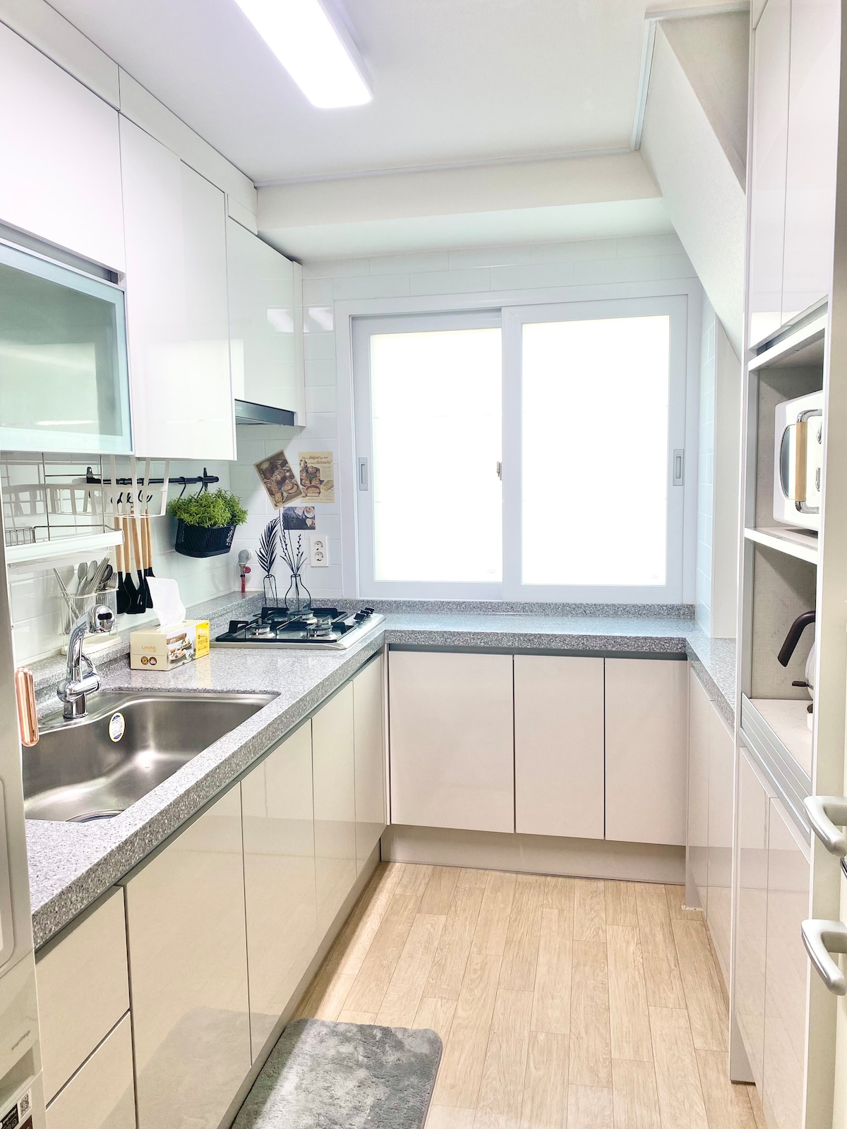 A well-equipped kitchen features sleek white cabinets and a polished countertop. Various appliances, including a stovetop and microwave, are available. Natural light floods in through a nearby window, enhancing the functional layout designed for convenience.