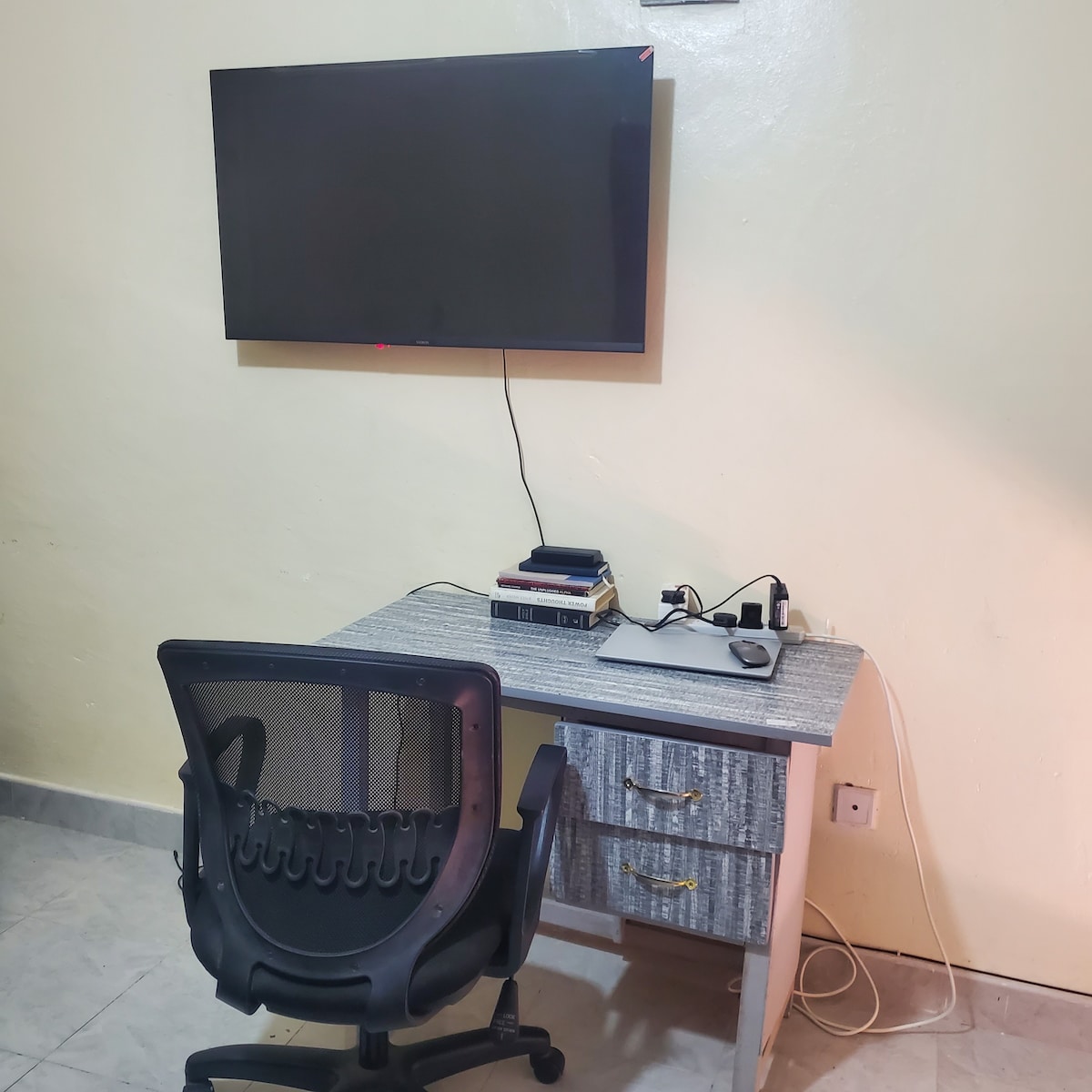 A workspace is depicted with a sleek desk featuring two drawers and a comfortable black office chair. A television is mounted on the wall above the desk, while a few books and electronic devices are neatly arranged on the surface, contributing to a functional atmosphere.