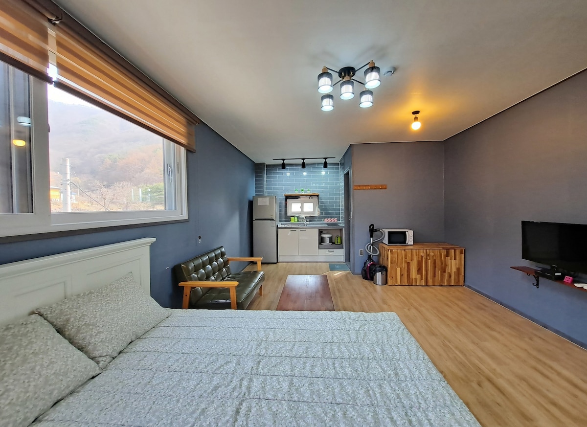 A spacious room features light wooden flooring and a large window providing natural light. A cozy sleeping area with a textured white bedspread is complemented by a sleek green sofa and wooden coffee table. The kitchenette, equipped with modern appliances, is visible in the background.