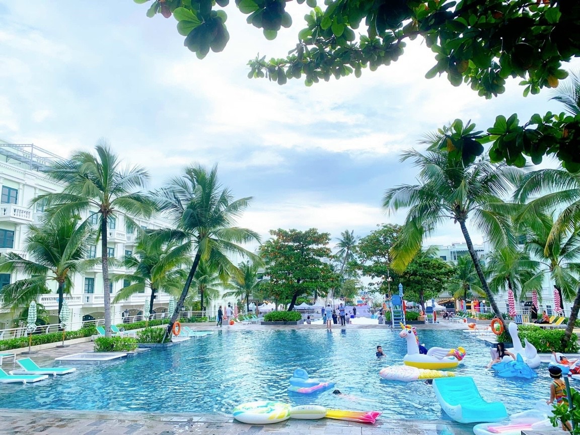 A large outdoor swimming pool is surrounded by palm trees, with floating inflatables scattered throughout the water. Lounge chairs are positioned along the pool's edge, and people can be seen enjoying the space, creating a relaxed atmosphere.