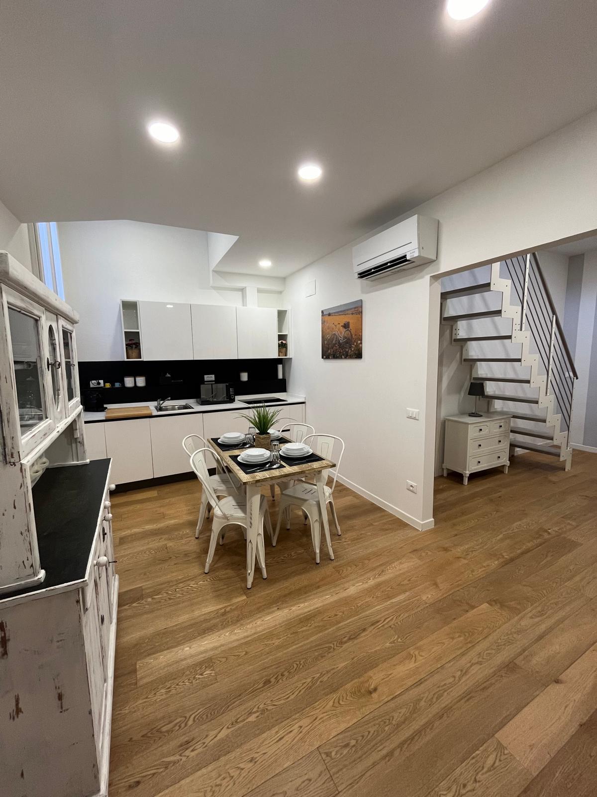 A modern kitchen area featuring white cabinetry and a dining table for four. The space is well-lit with recessed lighting, and a sleek black countertop contrasts with the light wooden flooring. Steps lead to an upper level, enhancing the open layout.