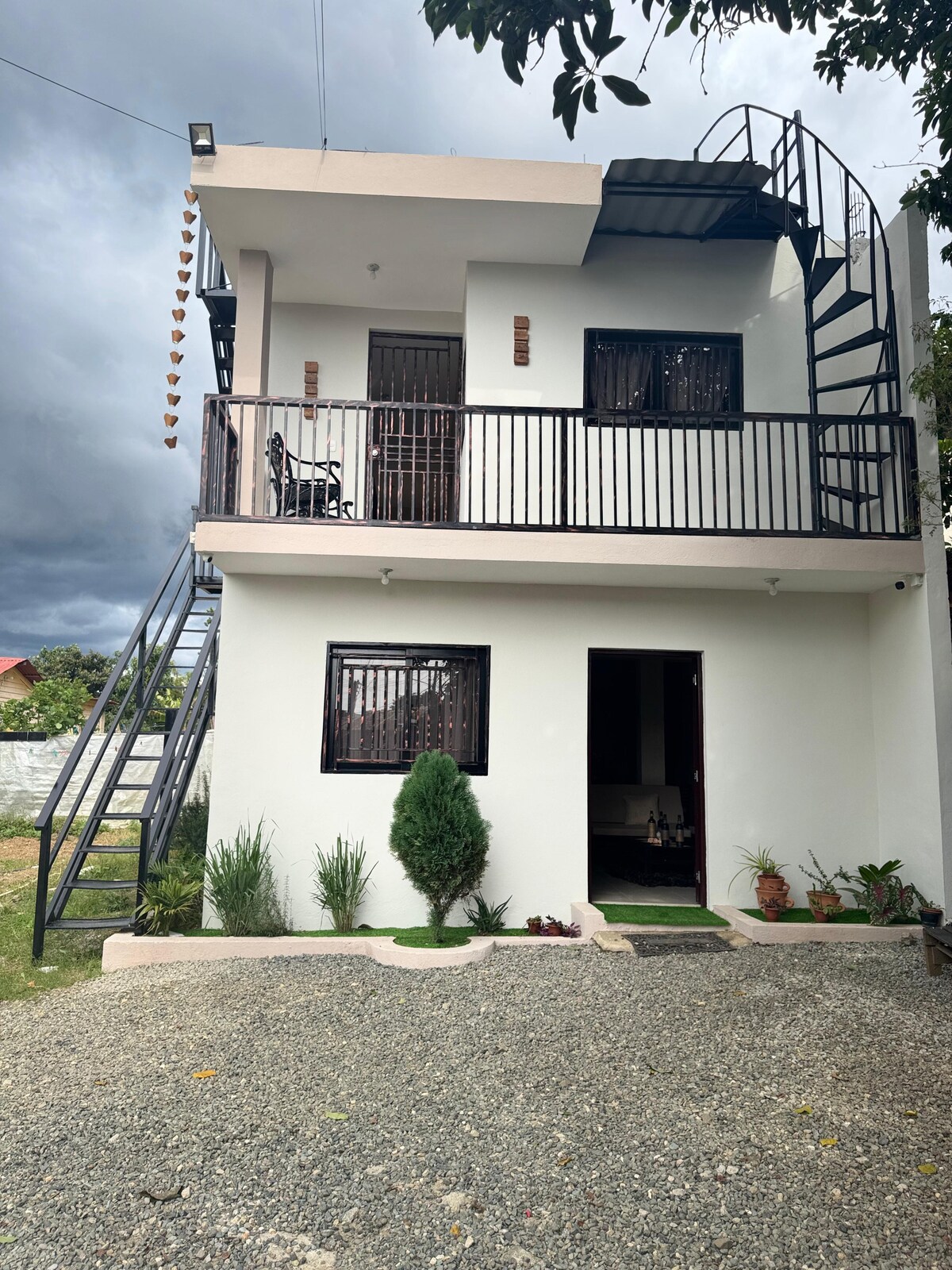 A two-story building is showcased with a modern design. A spiral staircase leads to a balcony featuring two chairs. The entrance is framed by small green plants and gravel paths, while large windows allow natural light to enter.