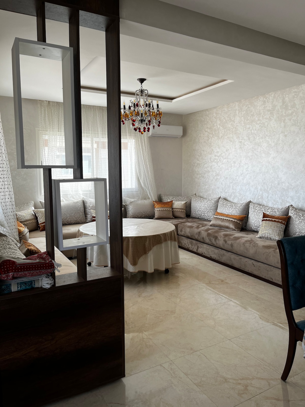 A well-appointed living area features a comfortable beige sofa with decorative cushions, arranged around a circular table. Light filters through sheer curtains, highlighting a stylish chandelier overhead. The refined decor is complemented by elegant marble flooring and a glimpse of a partition with modern geometric design.