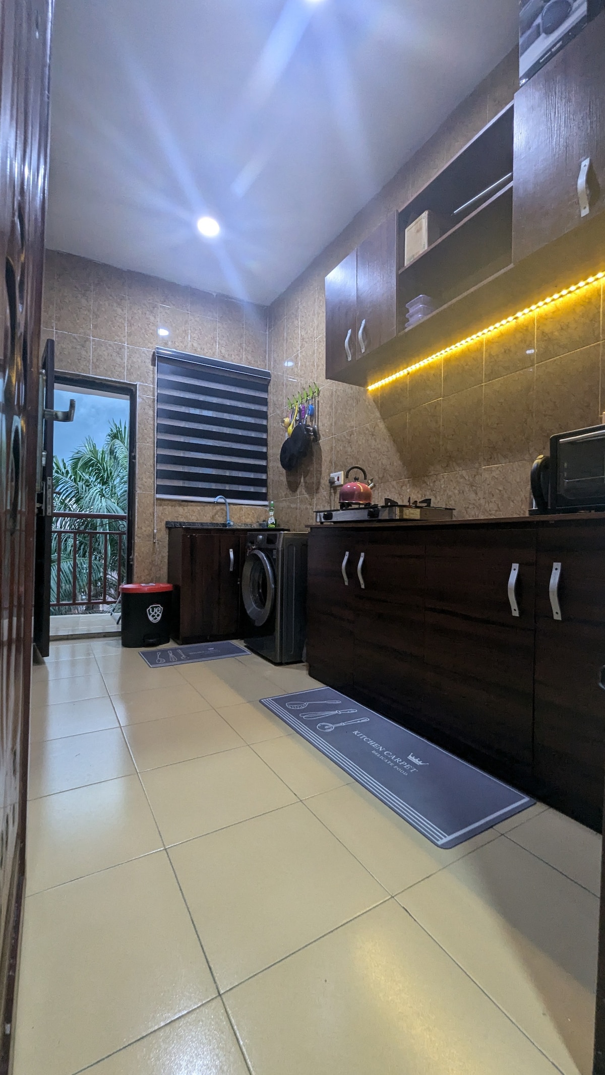 A compact kitchen area features modern wooden cabinetry and a variety of appliances, including a washing machine and microwave. Soft lighting highlights the countertops, while a window offers views of greenery outside. A practical floor mat adds comfort beneath. Neat organization is visible throughout.
