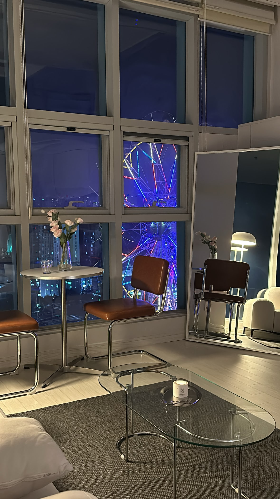 A cozy seating area is highlighted by two modern chairs at a small round table. A stylish mirror reflects the room's ambiance, while vibrant colors from a nearby Ferris wheel illuminate the night view through expansive windows.