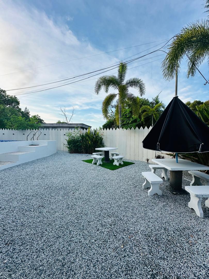 An outdoor space is visible, featuring a gravel surface with scattered greenery. Two white picnic tables are placed on a patch of grass, accompanied by white benches. A large black umbrella provides shade, with a white fence delineating the area.
