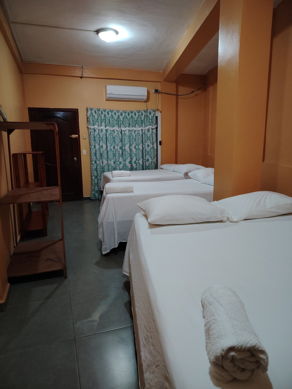 A cozy sleeping area features three spacious beds arranged with white linens and neatly folded towels. Walls are painted a warm orange, complemented by a teal patterned curtain covering a window. A wooden shelf stands against one wall, adding functional space.