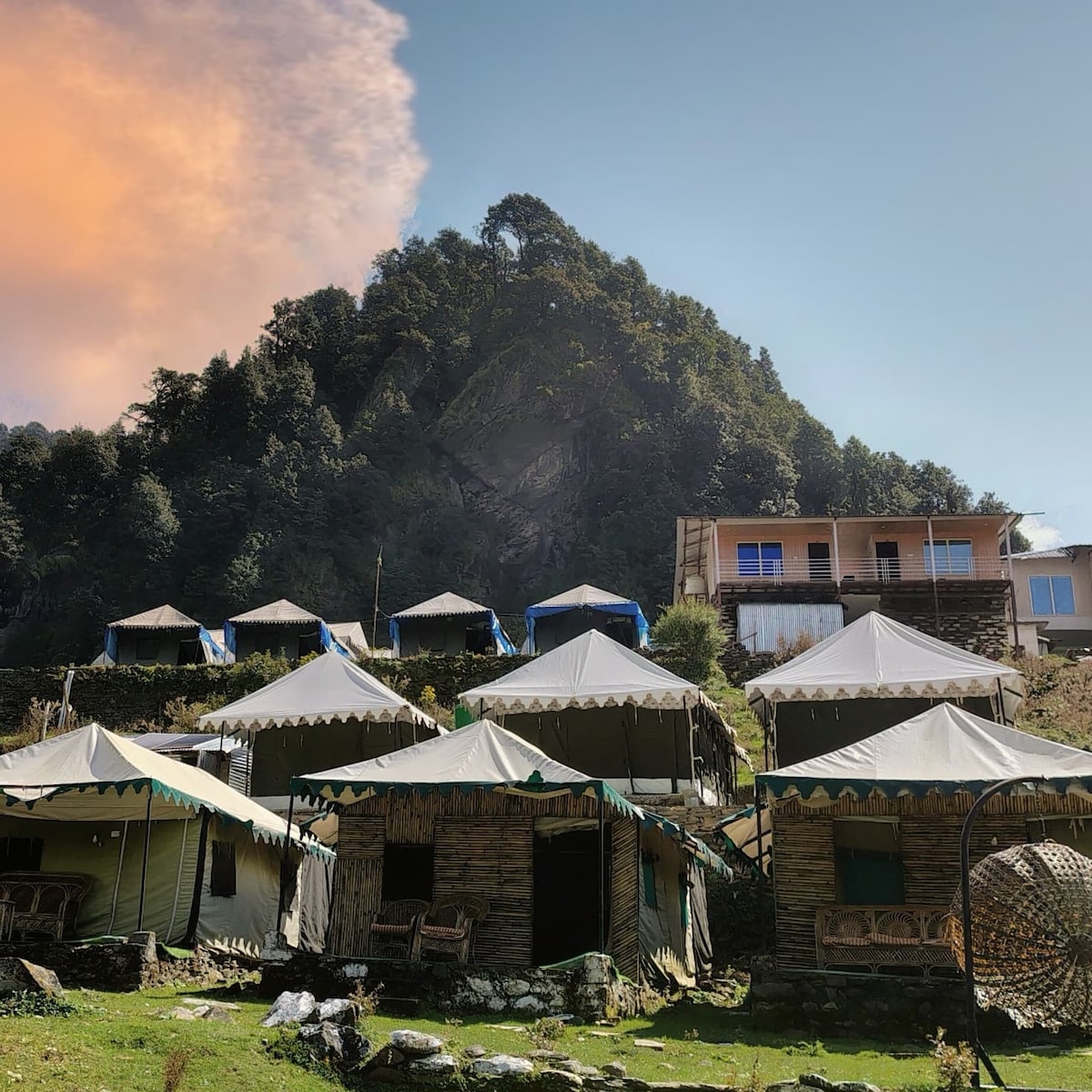A series of rustic tents is set against a lush green hillside, each featuring a canopy and wooden accents. In the background, a two-story building is visible, surrounded by trees and mountains. The sky displays a gradient of warm colors towards the top.