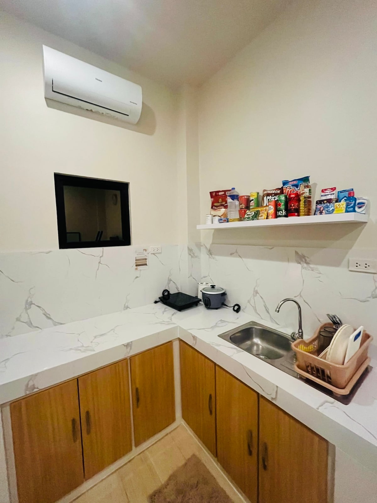 A compact kitchen area features modern cabinetry in warm wood tones. A marble-patterned countertop houses a sink, cooking appliances, and utensils. A shelf displays a variety of packaged food items. An air conditioning unit is mounted on the wall, providing comfort.
