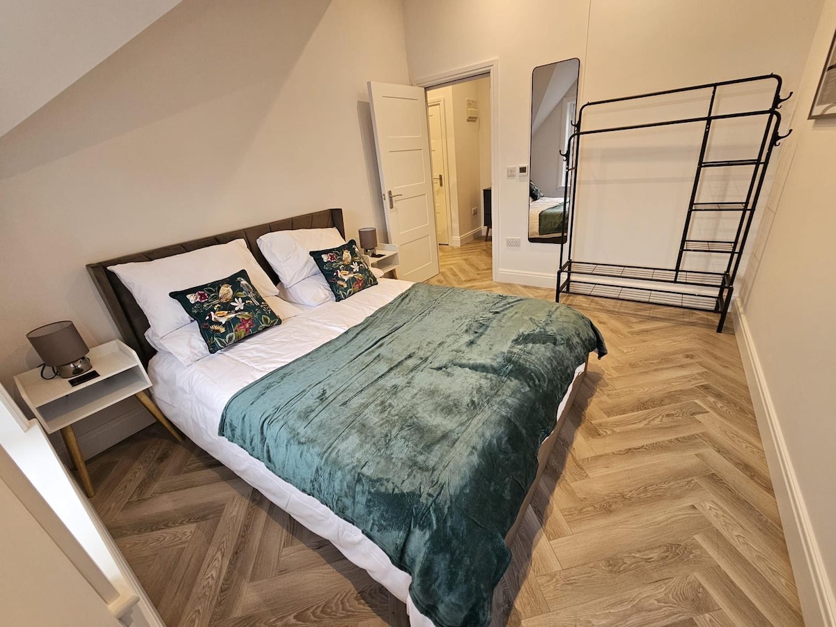 The bedroom features a spacious bed topped with a plush dark green throw and decorative pillows. A full-length mirror is positioned beside a clothing rack, while warm, natural light enhances the light-colored walls and wooden flooring.