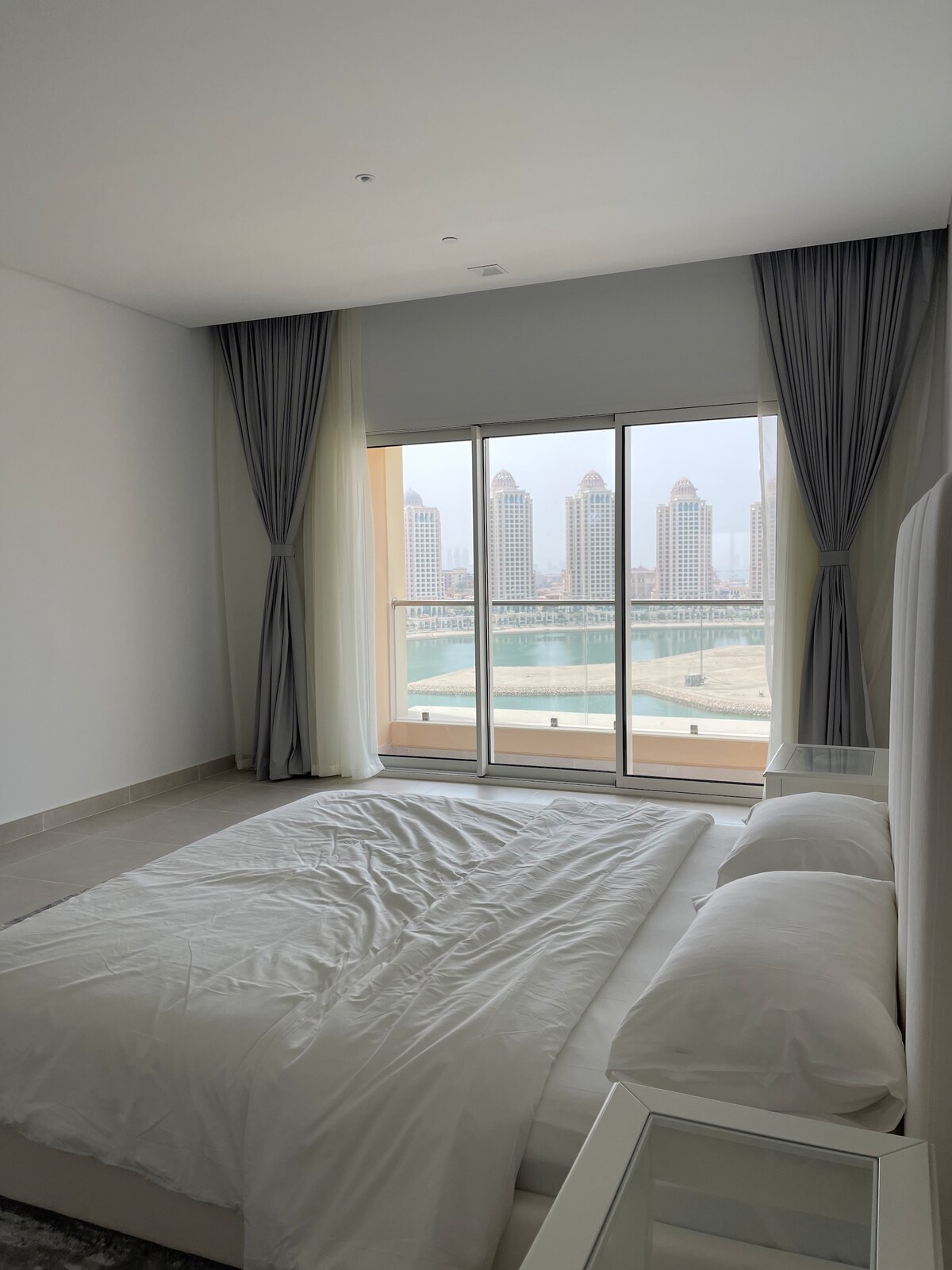 A spacious bedroom features a large bed with white bedding, complemented by a nightstand. Floor-to-ceiling windows allow ample natural light, offering a view of the surrounding buildings and sea. Elegant curtains frame the windows, enhancing the room's serene atmosphere.