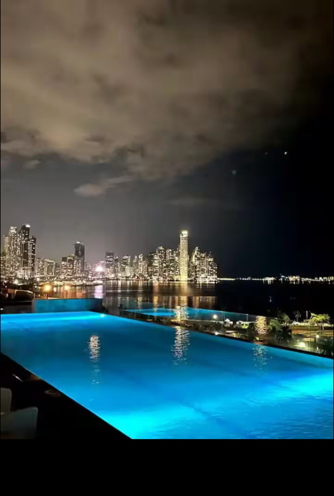 An infinity pool is illuminated with blue lighting, offering a view of a vibrant city skyline at night. The buildings reflect off the water's surface, while soft clouds drift across the sky, contributing to the serene atmosphere.