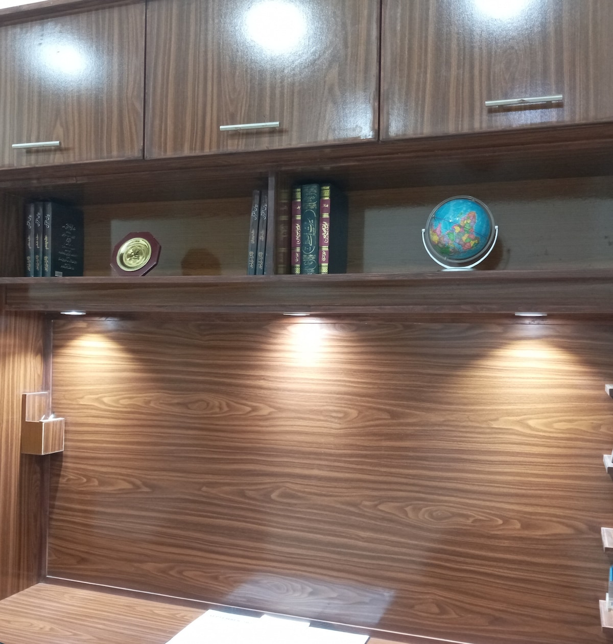 A study area is shown with a polished wooden desk and shelving. Below, soft lighting illuminates the desktop, while above, neatly arranged books and decorative items, including a globe and plaques, complement the organized appearance.