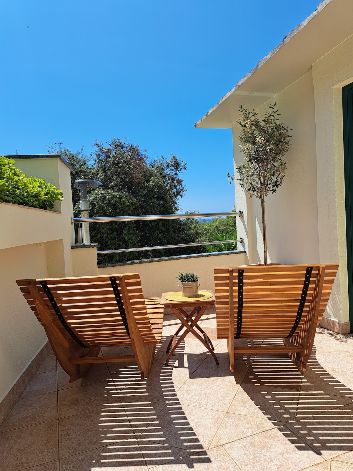 A sunny terrace features two wooden deck chairs arranged facing a small round table. A potted olive tree adds a touch of greenery, while the surrounding walls provide privacy. A clear blue sky and distant foliage are visible, enhancing the tranquil outdoor setting.