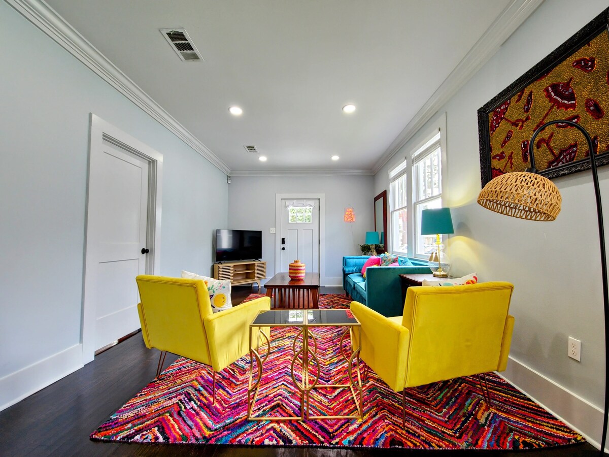 A colorful living room features vibrant yellow chairs paired with a teal sofa. A coffee table sits on a patterned rug, and a television is positioned against the wall. Natural light floods the space through large windows, highlighting the eclectic decor.