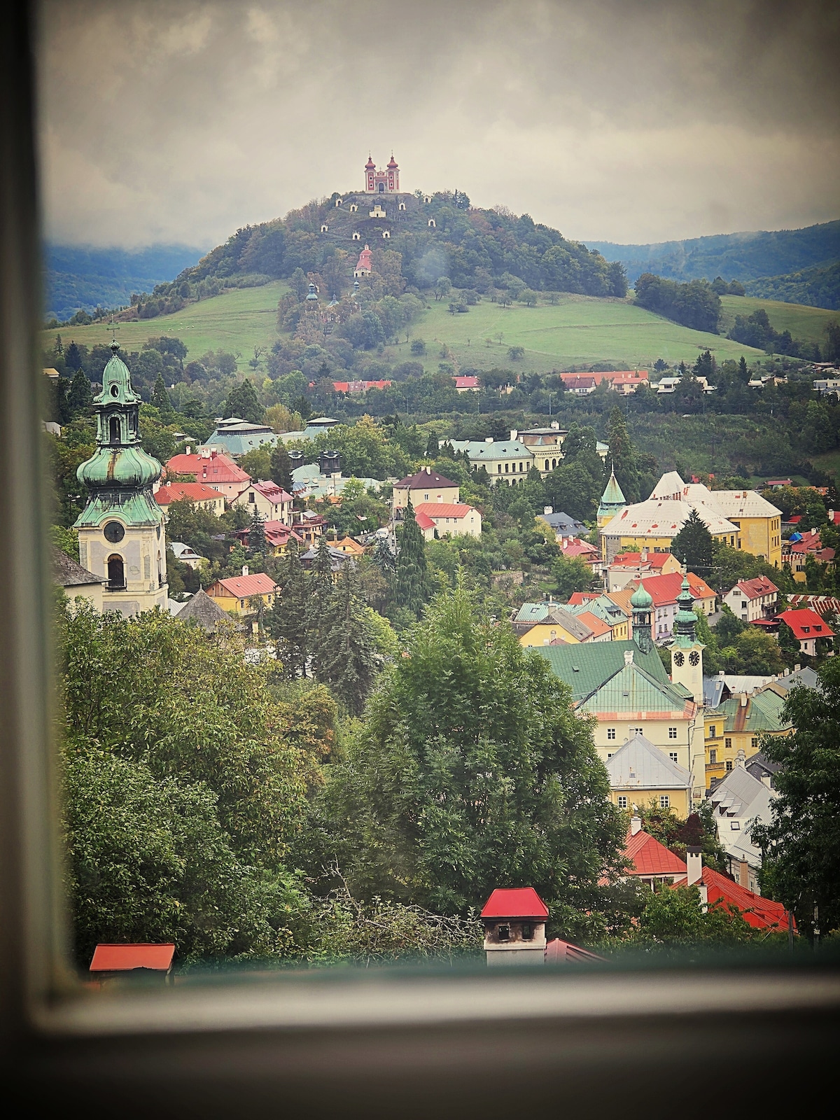 A scenic view is captured from a window, showcasing the charming rooftops of Banská Štiavnica. Rolling green hills are visible in the background, with a historic church steeple in the foreground and the prominent hill featuring a castle at the top.