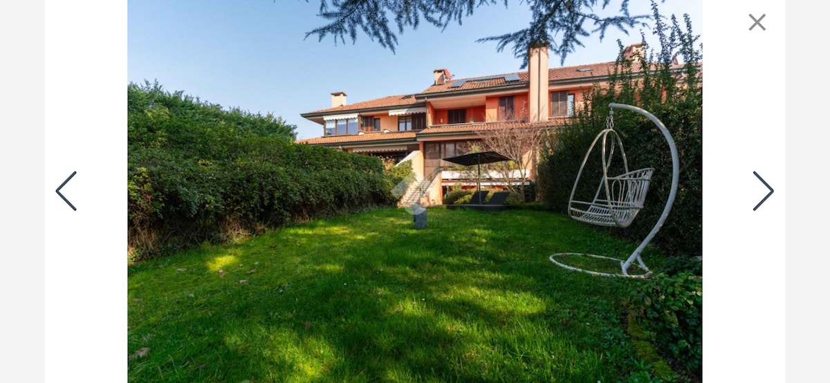 A lush green garden is featured, with a comfortable hanging chair positioned on the grass. The backdrop includes a charming multistory building, partially obscured by well-maintained hedges. Clear skies enhance the soothing ambiance of this outdoor space.