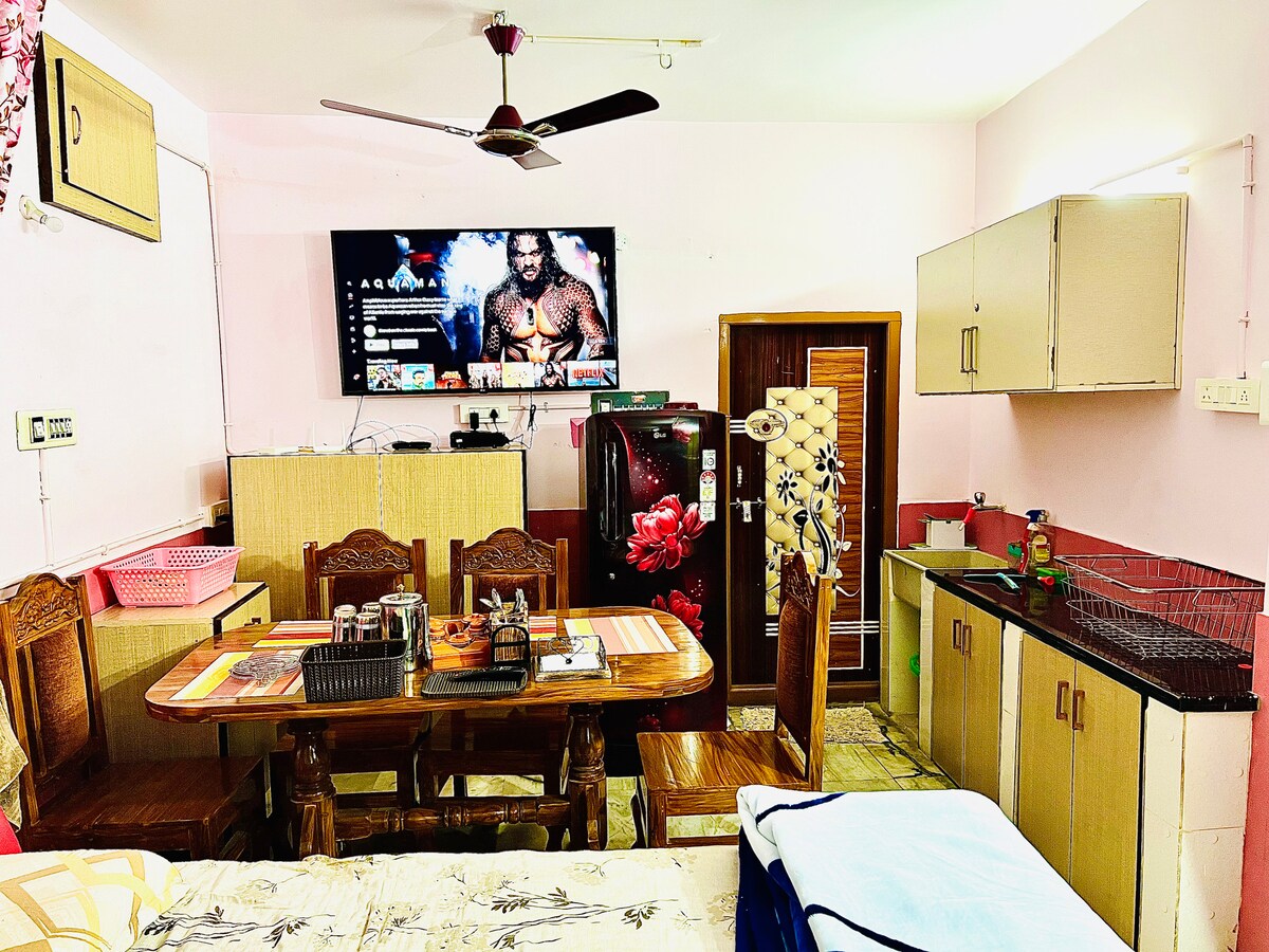 A cozy living area features a dining table surrounded by wooden chairs and a wall-mounted television. The kitchen area includes cabinetry and a sink, complemented by decorative elements. Soft lighting enhances the warm atmosphere, creating a welcoming space for guests.
