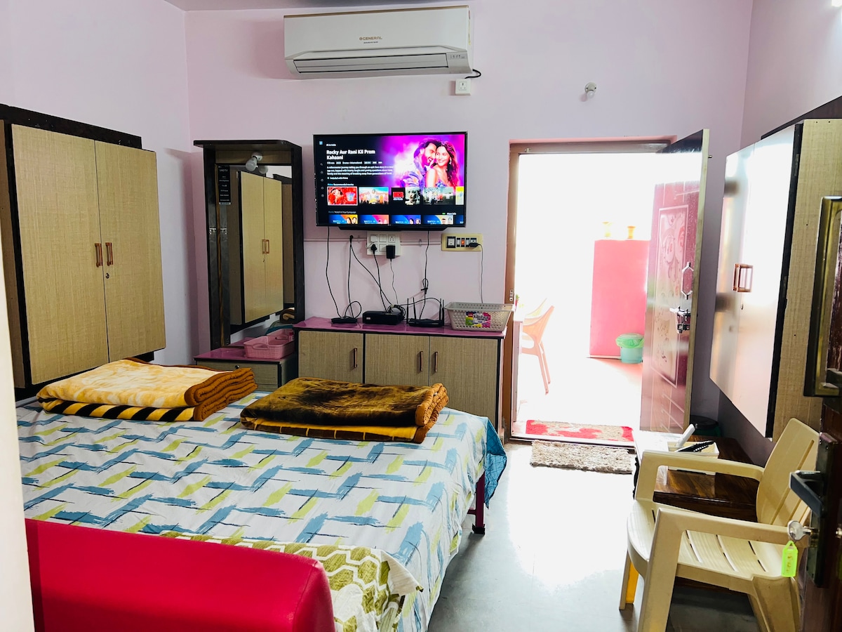 A well-furnished room is presented, featuring a spacious bed covered with a patterned blanket. A large wall-mounted television displays vibrant content. Cabinets line the walls, and a chair is positioned nearby. A doorway leads to an outdoor area, enhancing accessibility.