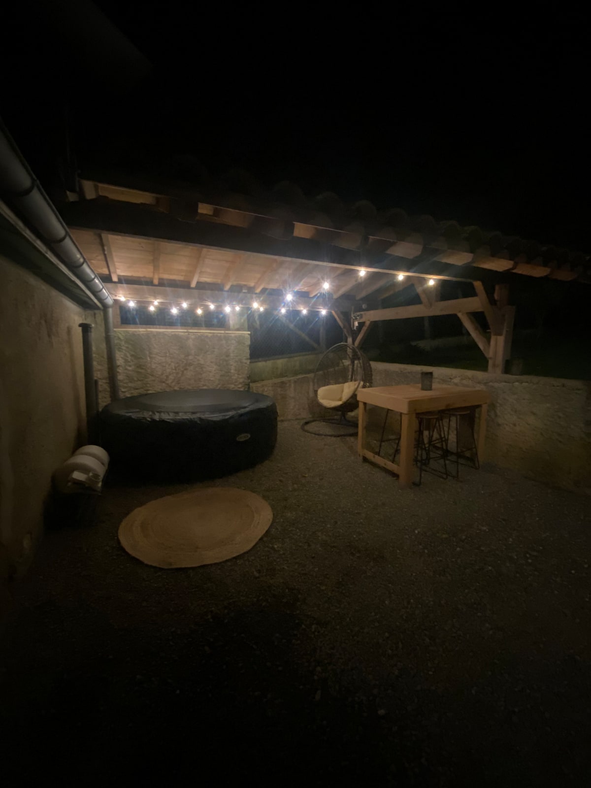 A covered terrace is illuminated by soft string lights, creating a cozy atmosphere. A round black bean bag sits next to a sturdy wooden table, complemented by a hanging chair and a soft circular rug on the gravel floor.
